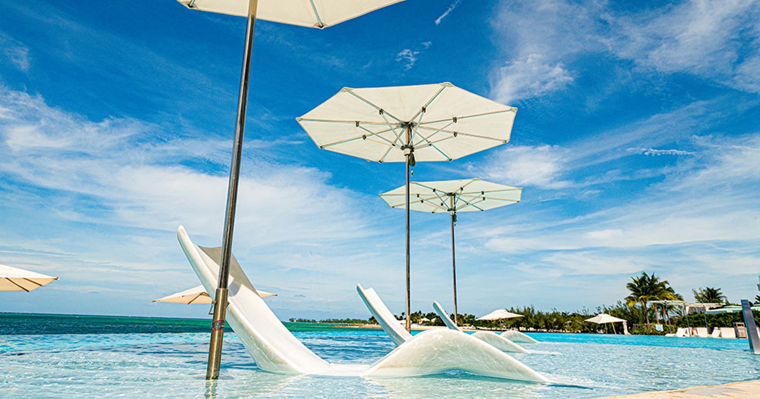 White beach chairs and umbrellas by a pool with blue sky and ocean in the background