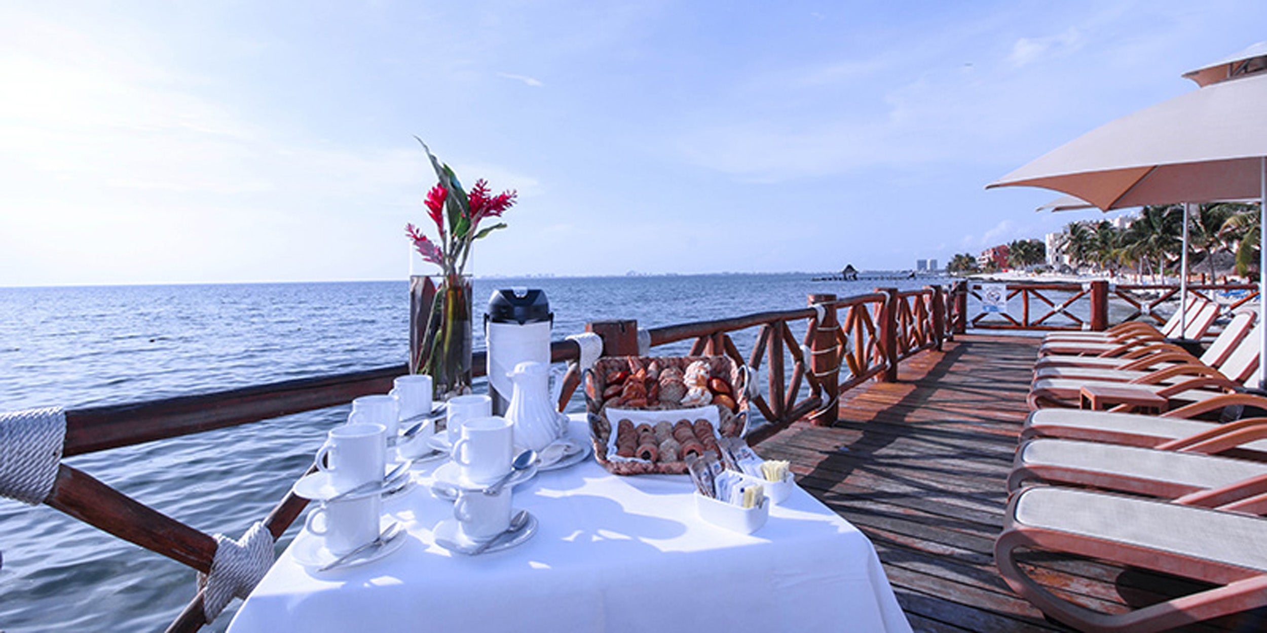Dining table set for two on a deck by the ocean with chairs and umbrellas.