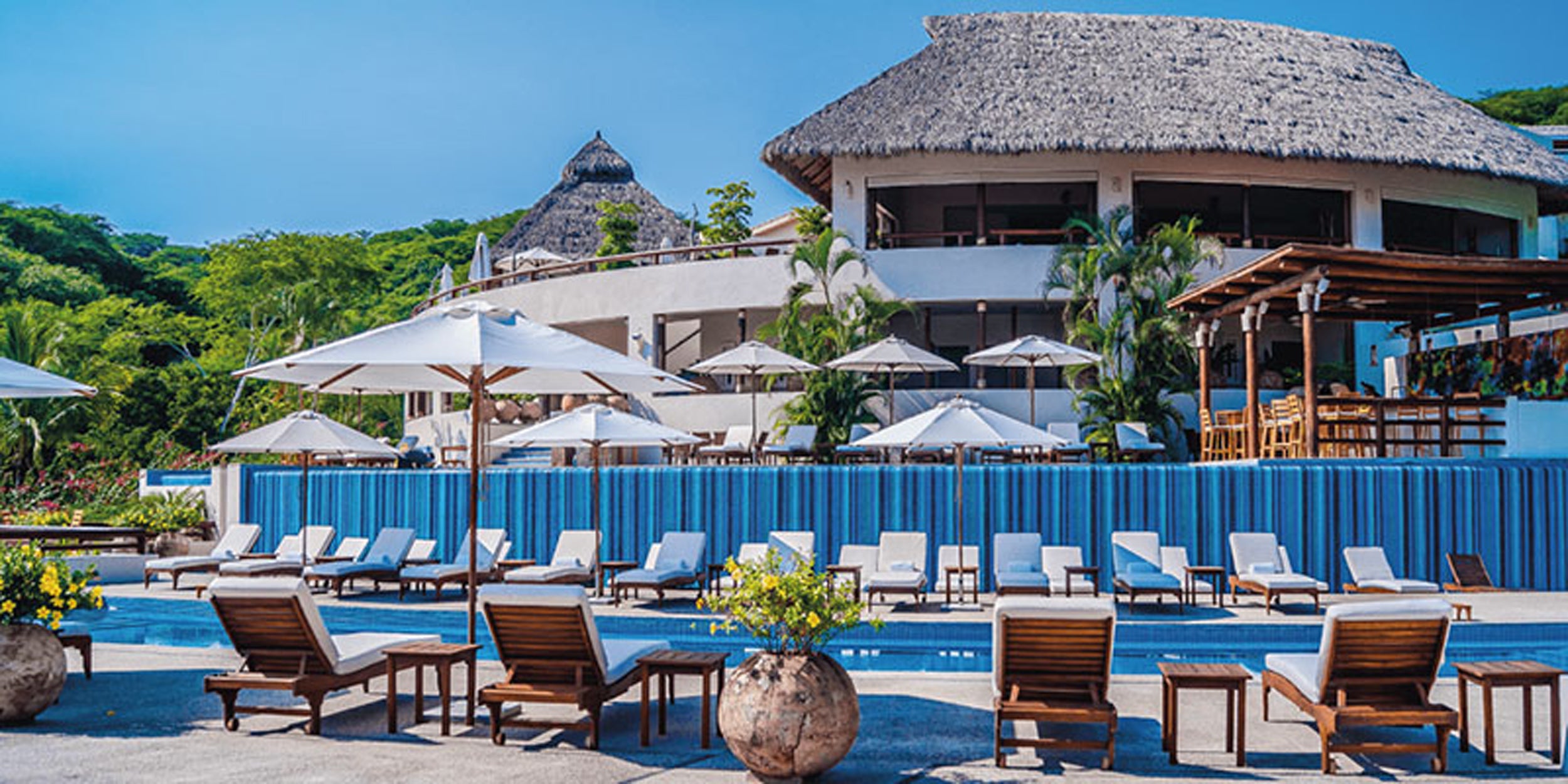 Pool area with lounge chairs, umbrellas, and a building with a thatched roof in the background.