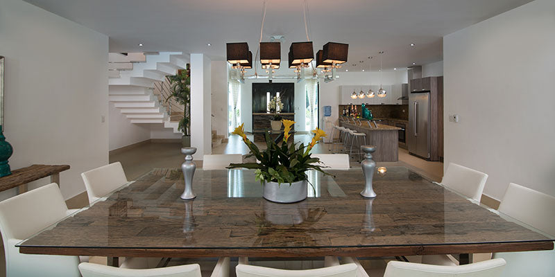 Modern dining room with a long wooden table and chairs, featuring a kitchen in the background.