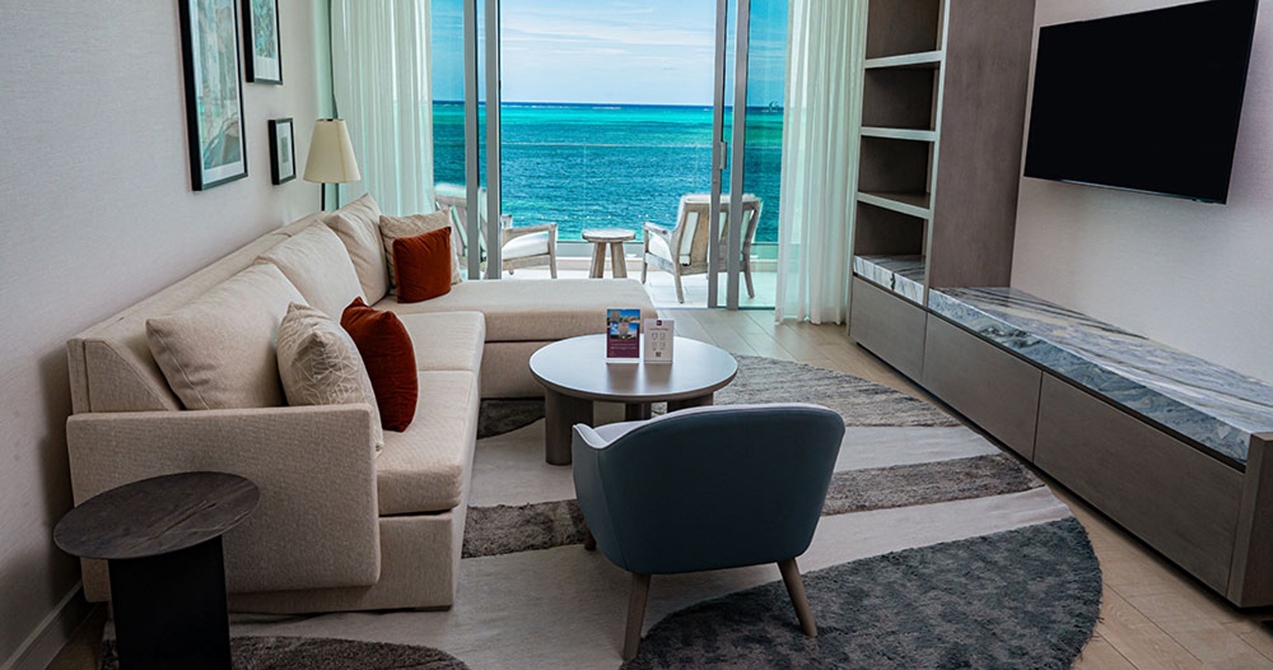 Modern living room with a view of the ocean, featuring a sofa, chairs, and a television.