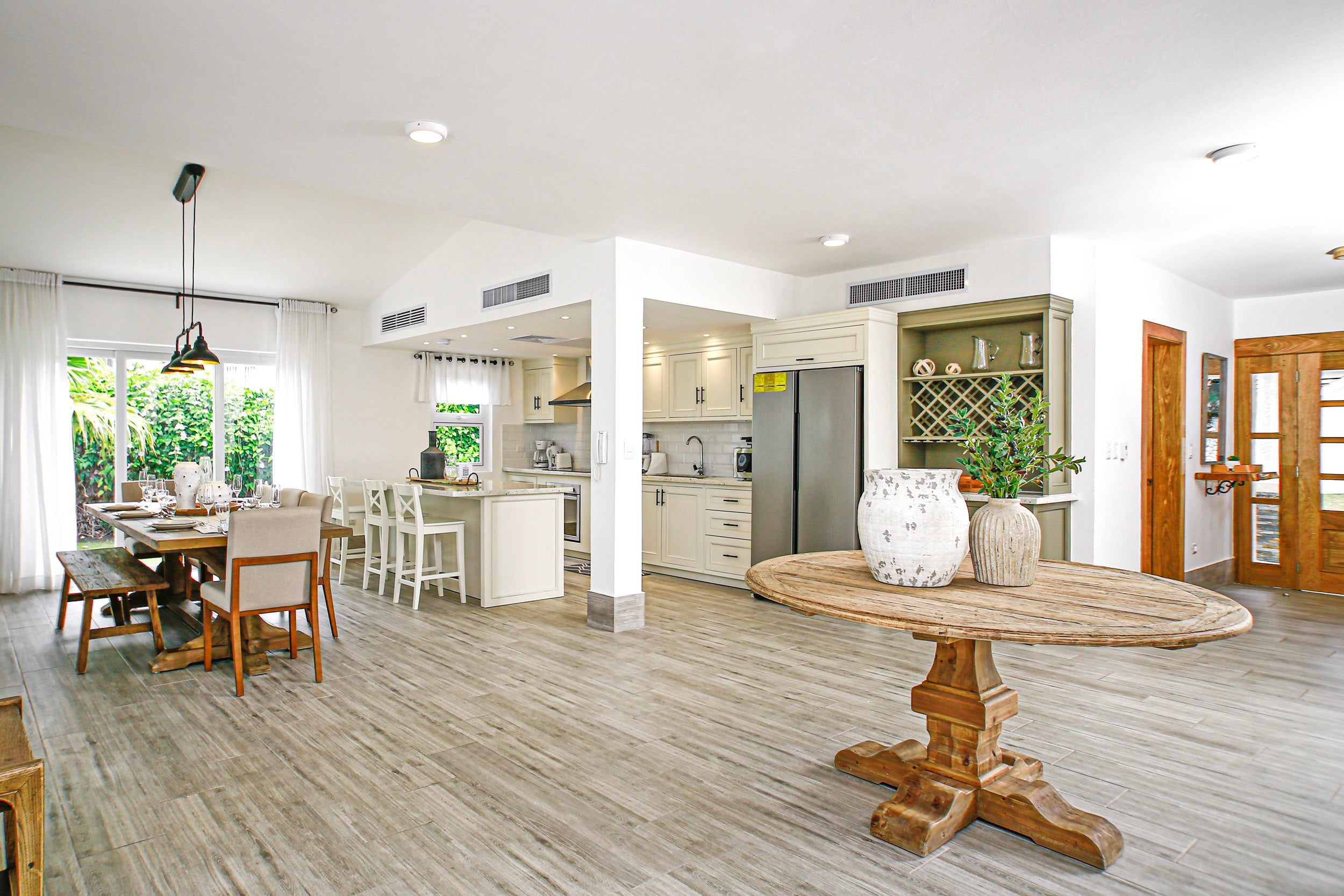 Open-plan kitchen and dining area with wooden table and chairs, white cabinets, and large windows.