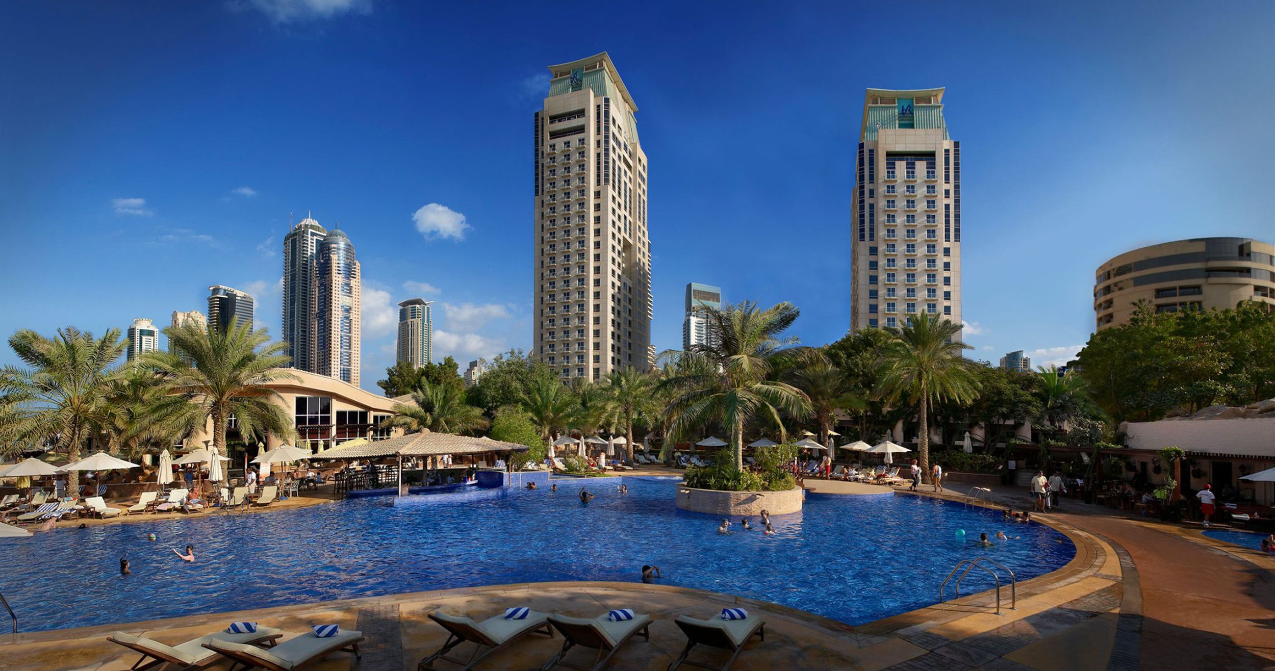 Pool area with lounge chairs and palm trees, surrounded by tall buildings under a clear blue sky.