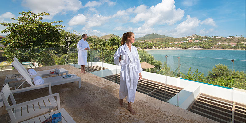 Two people in white robes standing on a terrace with a scenic view of the ocean and mountains.